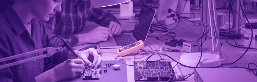 Team of engineers working on electronic components in a lab with monitors displaying schematics.